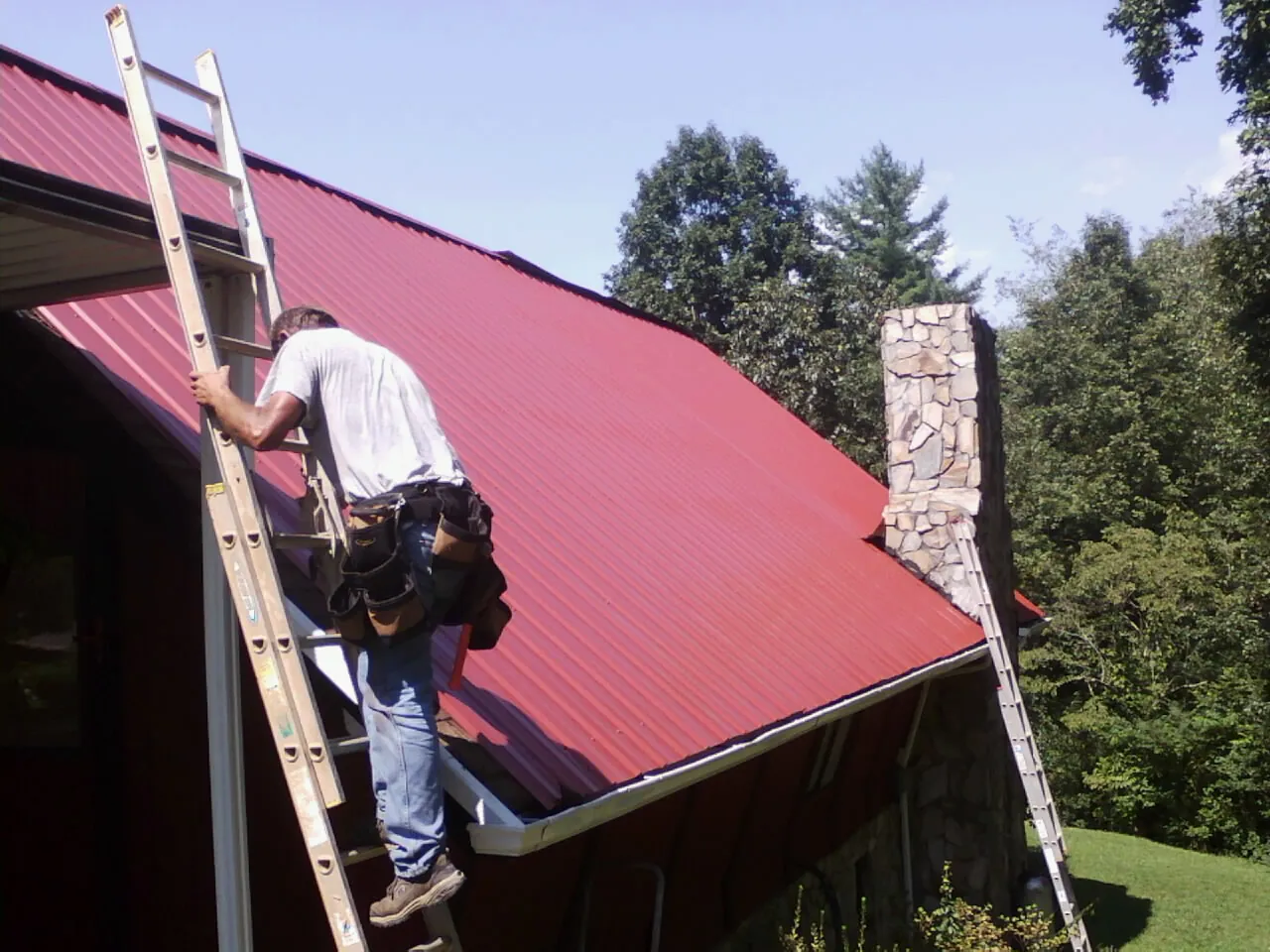 Roofer on a ladder beside a red metal roof and stone chimney — Soffit & Fascia Repair by Roofing Brothers in Lowgap